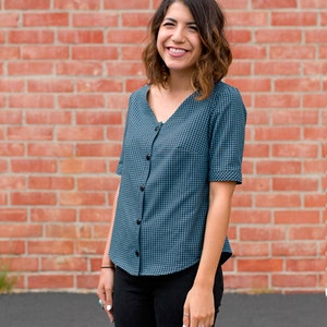 May include: A teal and black gingham button-down shirt with short sleeves, a V-neckline, and a curved hemline. The shirt features a series of small black buttons running down the center front, paired with solid black trousers.