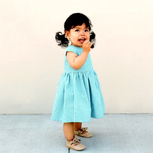 May include: A toddler dress featuring a teal and white gingham check pattern with a round neckline, cap sleeves, and a gathered skirt. The dress is paired with beige leather-style shoes that have a small bow detail on the front of each shoe.