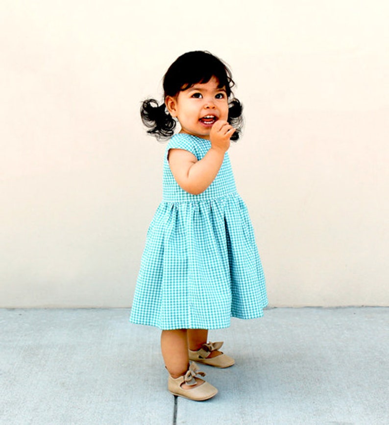May include: A toddler dress featuring a teal and white gingham check pattern with a round neckline, cap sleeves, and a gathered skirt. The dress is paired with beige leather-style shoes that have a small bow detail on the front of each shoe.