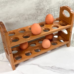 May include: A two-tiered wooden egg holder with 24 individual slots. The holder is made of light brown wood and has a rustic look. There are five brown eggs in the holder.
