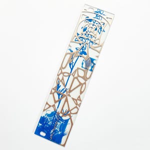 May include: A rectangular bookmark with a white background, featuring blue floral and geometric patterns. The bookmark has a laser-cut, copper-colored overlay with intricate designs. The bottom section has a blue illustration. The bookmark is approximately 7 inches long.