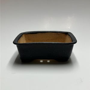 May include: A black rectangular bonsai pot with a brown interior. The pot has a textured surface and three drainage holes.