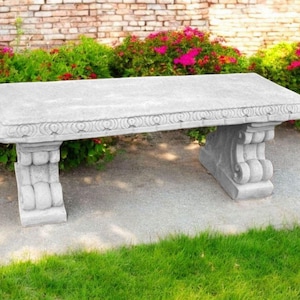 May include: A white stone bench with ornate detailing sits on a gray stone path surrounded by green grass and pink flowers.