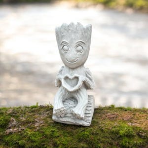 May include: A gray statue of a character holding a heart-shaped symbol. The statue has a smiling face and is seated on a rectangular base. The statue is made of a stone-like material and is placed on a bed of green moss.