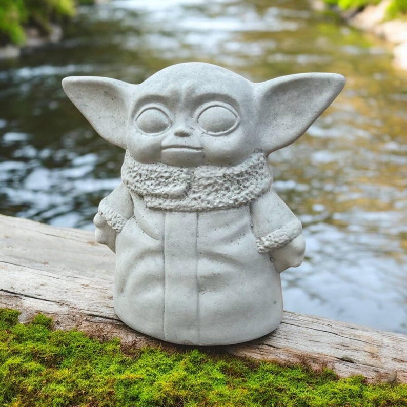 K&ouml;nnte beinhalten: Eine graue Betonstatue einer Figur mit gro&szlig;en Ohren und einem Kapuzenmantel. Die Statue steht auf einem Holzstamm mit gr&uuml;nem Moos vor einem verschwommenen Hintergrund aus Wasser und Gr&uuml;n.