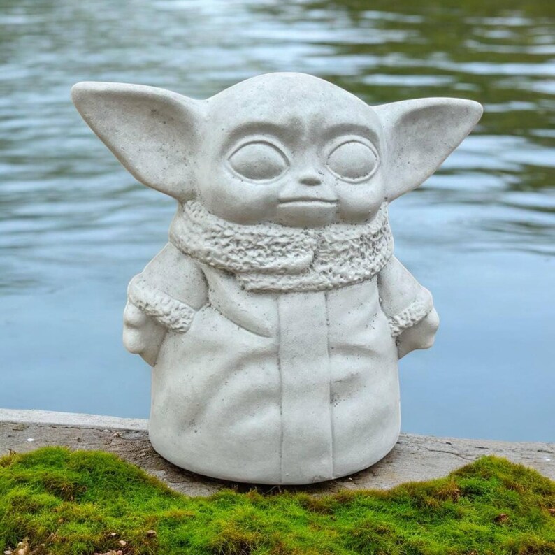 K&ouml;nnte beinhalten: Eine hellgraue Statue einer Figur mit gro&szlig;en Ohren und einem Kapuzenmantel. Die Statue hat einen detaillierten Schal und steht auf einer Holzoberfl&auml;che mit gr&uuml;nem Moos vor einem Gew&auml;sser. Die Statue ist eine beliebte Figur aus einem Science-Fiction-Franchise.
