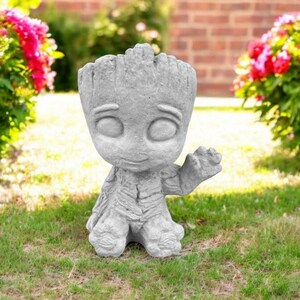 May include: A gray concrete garden statue of a character from the movie Guardians of the Galaxy. The statue is of Groot, a tree-like humanoid, with a friendly expression and one arm raised in a wave. The statue is standing on a grassy lawn.
