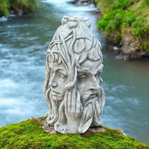 May include: A gray stone sculpture of two faces, one feminine and one masculine, with intertwined hair and foliage. The sculpture is set on a mossy surface with a river in the background. The faces are detailed with closed eyes and hands near the face.