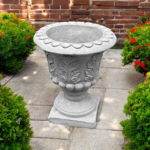May include: A gray concrete planter with a decorative leaf design. The planter is sitting on a stone patio surrounded by green bushes.