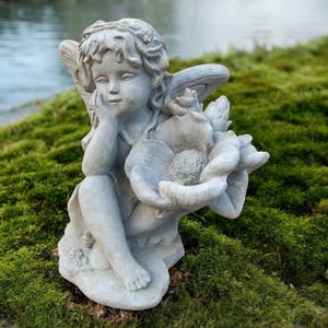 May include: A gray stone angel statue, seated and holding a large flower. The angel has closed eyes and rests its chin on its hand. The statue is set on a bed of green moss, with a body of water in the background.