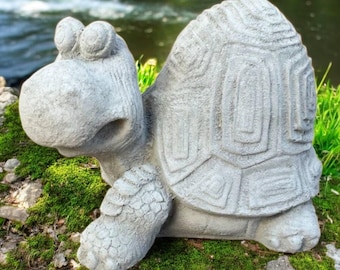 May include: A whimsical, gray concrete turtle statue with a detailed shell pattern and a smiling face. The garden ornament is placed on a mossy rock, with a blurred river in the background. The turtle has large, expressive eyes and a textured surface.