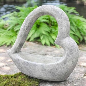 May include: A gray, heart-shaped garden planter made of stone. The planter has a curved handle and a shallow basin for holding plants or flowers. The textured surface gives it a rustic appearance. It is sitting on a stone surface.
