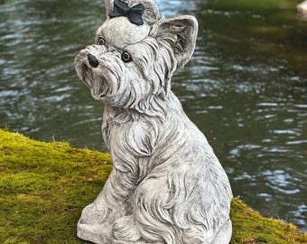 May include: A detailed, gray stone statue of a seated dog with a black bow on its head. The dog has a textured coat and is positioned on a bed of green moss, with a river in the background. The statue is a decorative garden ornament.