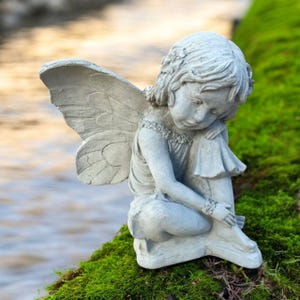 May include: A gray concrete fairy statue with detailed wings, seated on a bed of green moss. The fairy is depicted with a serene expression, her head resting on her knees. The statue is positioned near a body of water.