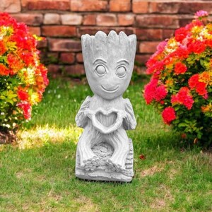 May include: A gray concrete garden statue of a character holding a heart. The statue is sitting on a bed of green grass.