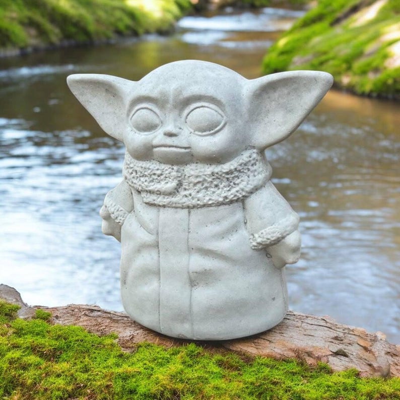 K&ouml;nnte beinhalten: Graue Betonstatue einer Figur mit gro&szlig;en Ohren und einem Kapuzenmantel. Die Statue steht auf einem St&uuml;ck Holz, das mit gr&uuml;nem Moos bedeckt ist, mit einem verschwommenen Hintergrund aus Bach und Gr&uuml;n.