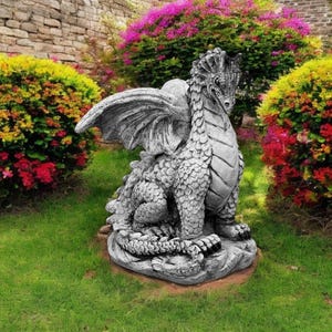 May include: A gray stone dragon statue with wings outstretched, sitting on a rock base in a garden setting. The dragon has a detailed, scaly texture and a fierce expression.