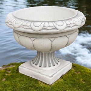 May include: A large, off-white, ornate planter with a wide, scalloped rim. The planter has a bulbous body with vertical grooves and sits on a square base. The background features water and greenery, suggesting an outdoor setting.