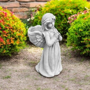 May include: A gray concrete statue of an angel with wings, wearing a long dress and holding her hands together in prayer. The statue is standing on a bed of brown grass.