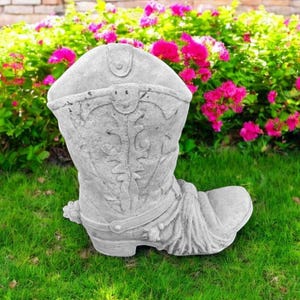 May include: A gray concrete cowboy boot planter with a decorative design. The boot is sitting on a green grassy surface.