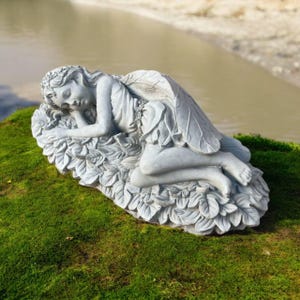 May include: A gray stone garden statue of a sleeping fairy with detailed wings and a floral crown. The fairy is lying on a bed of leaves, creating a serene and whimsical outdoor decoration. The statue is placed on a bed of green moss.