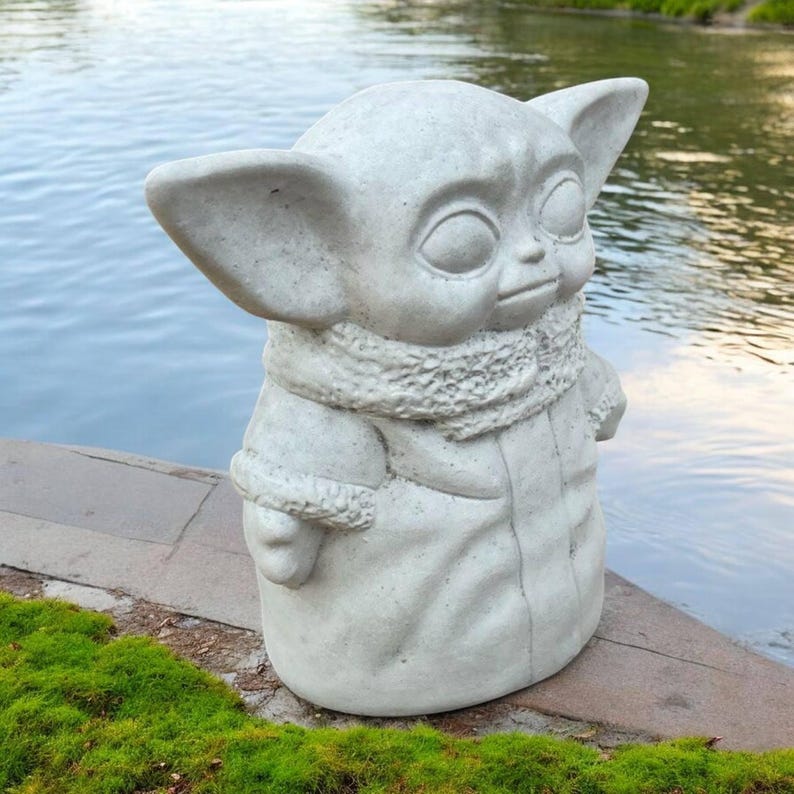 K&ouml;nnte beinhalten: Eine hellgraue Statue einer Figur mit gro&szlig;en Ohren, gro&szlig;en Augen und einem Schal. Die Statue steht auf einer Steinoberfl&auml;che neben einem Gew&auml;sser. Die Statue tr&auml;gt einen Mantel mit fell&auml;hnlichem Besatz.