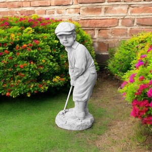 May include: A gray concrete statue of a golfer wearing a cap and holding a golf club. The statue is standing on a small base and is surrounded by green grass and colorful flowers.