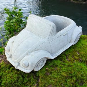 May include: A light gray concrete planter shaped like a classic convertible car. The planter has an open top for planting and sits on a bed of green moss. The car has detailed headlights, wheels, and door handles.