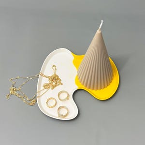 May include: A white and yellow ceramic tray with a brown cone-shaped candle and gold jewellery. The tray is shaped like a paint palette.