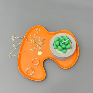 May include: An orange, kidney-shaped tray holds a gold necklace, two gold rings, and a small succulent-shaped candle in a concrete holder. The items are arranged on a grey background.