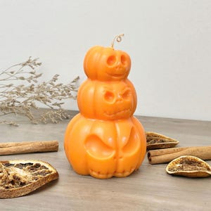 May include: A stacked pumpkin candle with three orange pumpkins, each with a carved jack-o'-lantern face. The candle is on a wooden surface with cinnamon sticks and dried orange slices.