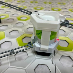 Puede incluir: Un componente de juguete de pl&aacute;stico blanco y transparente con un interior verde, conectado a pistas grises. El componente se encuentra sobre una superficie de baldosas hexagonales con detalles en verde y blanco. Una luz roja es visible en el componente.