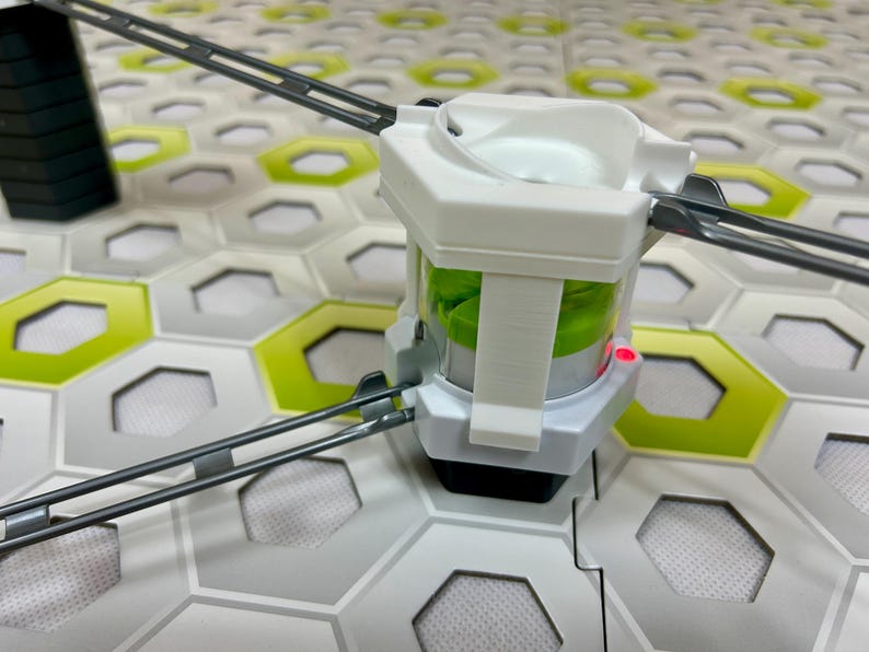 Puede incluir: Un componente de juguete de pl&aacute;stico blanco y transparente con un interior verde, conectado a pistas grises. El componente se encuentra sobre una superficie de baldosas hexagonales con detalles en verde y blanco. Una luz roja es visible en el componente.