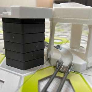 Puede incluir: Primer plano de un juego de construcci&oacute;n. El conjunto incluye una pila de bloques grises oscuros rematados con una pieza blanca, y una estructura blanca con una base curva y ra&iacute;les met&aacute;licos. La base est&aacute; sobre una superficie estampada.