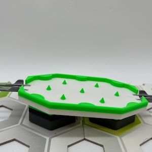 Pode incluir: Uma pe&ccedil;a de jogo de pl&aacute;stico branco e verde com pontas triangulares verdes. A pe&ccedil;a tem um centro branco e uma borda verde. Faz parte de um tabuleiro de jogo maior com ladrilhos hexagonais cinza e verdes.