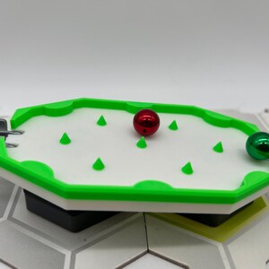 Pode incluir: Um pequeno tabuleiro de jogo com uma superf&iacute;cie branca, bordas verdes e formas c&ocirc;nicas verdes. Uma bola vermelha e uma verde met&aacute;lica est&atilde;o no tabuleiro. O tabuleiro faz parte de uma configura&ccedil;&atilde;o de jogo maior.