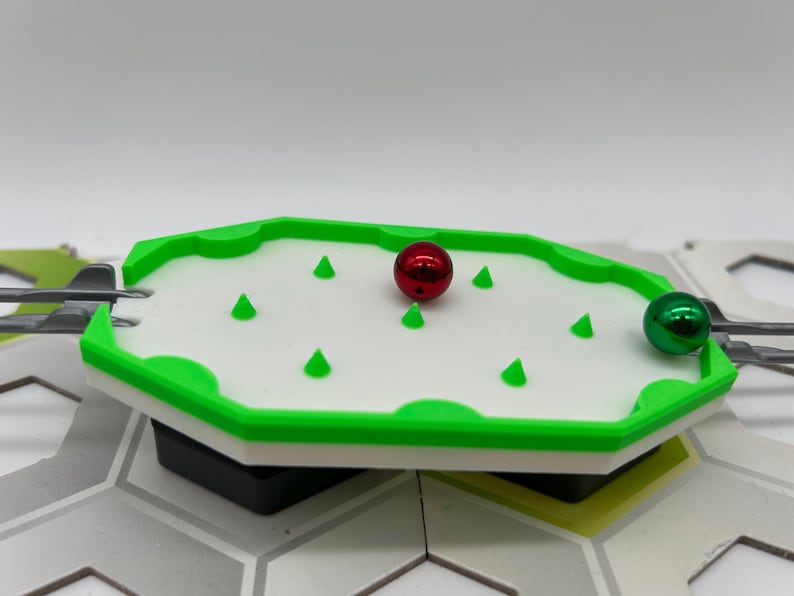 Pode incluir: Um pequeno tabuleiro de jogo com uma superf&iacute;cie branca, bordas verdes e formas c&ocirc;nicas verdes. Uma bola vermelha e uma verde met&aacute;lica est&atilde;o no tabuleiro. O tabuleiro faz parte de uma configura&ccedil;&atilde;o de jogo maior.