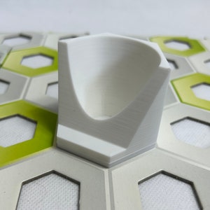 May include: A white plastic game piece shaped like a curved, open-top container. The piece is sitting on a hexagonal tile game board with green and gray accents.