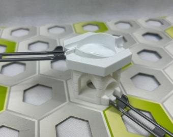 Puede incluir: Una torre de juguete de plástico blanco con una pista gris unida al lado. La torre tiene una abertura redonda en la parte superior y una abertura más pequeña en el lado. La torre está sentada en una baldosa hexagonal gris y verde.