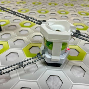 Puede incluir: Primer plano de una estructura de juguete blanca y gris con luces verdes y rojas. La estructura est&aacute; conectada a pistas grises y se encuentra sobre una superficie de baldosas hexagonales con detalles verdes y blancos. El juguete parece ser un circuito de canicas.