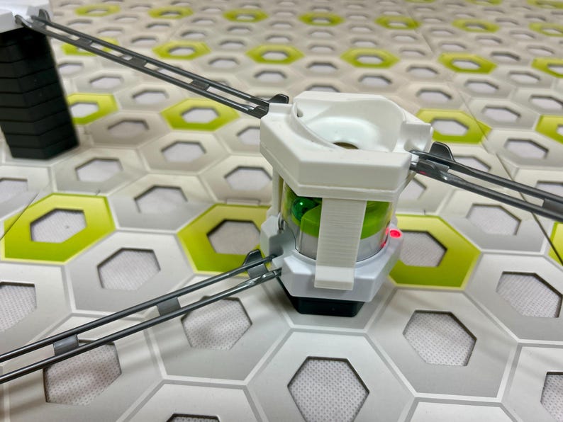 Puede incluir: Primer plano de una estructura de juguete blanca y gris con luces verdes y rojas. La estructura est&aacute; conectada a pistas grises y se encuentra sobre una superficie de baldosas hexagonales con detalles verdes y blancos. El juguete parece ser un circuito de canicas.