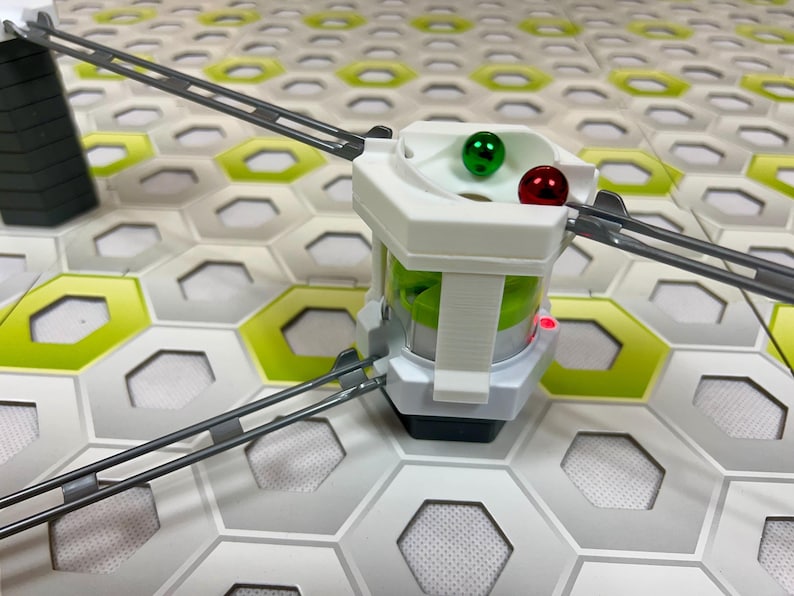 Puede incluir: Un juguete de circuito de canicas blanco y gris con una bola verde y otra roja en su interior. El juguete est&aacute; sobre una base de baldosas hexagonales con pistas grises que se extienden desde la estructura central. El juguete tiene un dise&ntilde;o futurista.