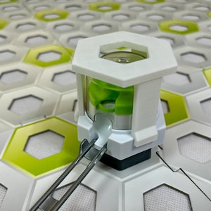 Puede incluir: Un dispositivo blanco y verde con una parte superior hexagonal y un interior cil&iacute;ndrico transparente. El dispositivo tiene un componente verde en el interior y est&aacute; conectado a una pista de metal plateado. El fondo presenta una superficie estampada con baldosas hexagonales blancas, verdes y grises.