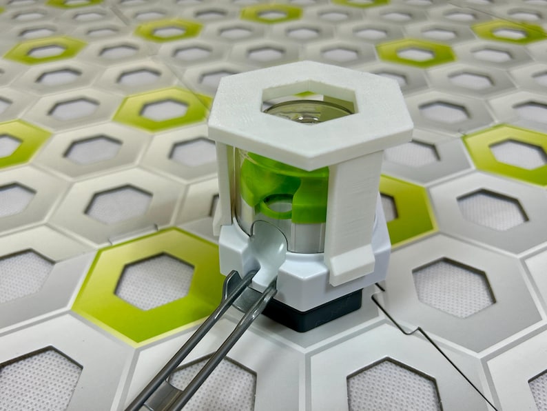 Puede incluir: Un dispositivo blanco y verde con una parte superior hexagonal y un interior cil&iacute;ndrico transparente. El dispositivo tiene un componente verde en el interior y est&aacute; conectado a una pista de metal plateado. El fondo presenta una superficie estampada con baldosas hexagonales blancas, verdes y grises.