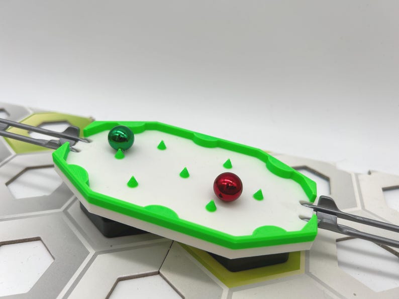 Pode incluir: Um tabuleiro de jogo branco e verde com uma borda verde e formas c&ocirc;nicas verdes. Duas esferas met&aacute;licas, uma verde e outra vermelha, est&atilde;o no tabuleiro. O tabuleiro faz parte de uma estrutura maior com formas geom&eacute;tricas cinza e brancas.