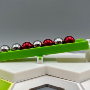 Könnte beinhalten: Ein grünes und weißes Spielzeug aus Plastik mit einer Rampe und einer Reihe von roten und silbernen Kugeln. Die Rampe ist nach oben geneigt und die Kugeln rollen die Rampe herunter.