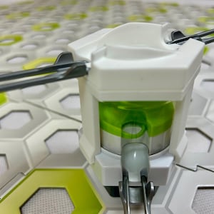 Puede incluir: Primer plano de un componente de juguete blanco y verde con varillas met&aacute;licas que se extienden hacia afuera. El componente tiene un interior cil&iacute;ndrico verde y est&aacute; colocado sobre un fondo de formas hexagonales blancas y verdes.