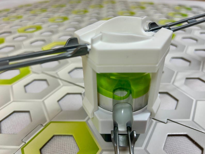 Puede incluir: Primer plano de un componente de juguete blanco y verde con varillas met&aacute;licas que se extienden hacia afuera. El componente tiene un interior cil&iacute;ndrico verde y est&aacute; colocado sobre un fondo de formas hexagonales blancas y verdes.