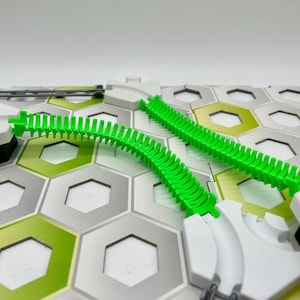 Op de afbeelding: Een groen baanstuk voor een knikkerbaan spel. De baan is flexibel en kan in verschillende vormen worden gebogen. De baan ligt op een wit en grijs zeshoekig speelbord.