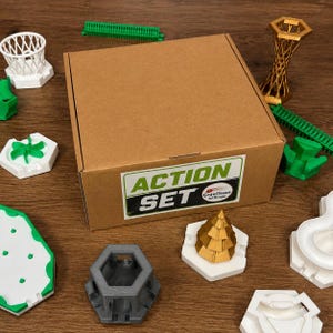 Può includere: Una scatola di cartone marrone con l'etichetta ACTION SET e il logo GraviTeam 3D Drucke, circondata da vari componenti stampati in 3D, tra cui un canestro da basket bianco, una torre dorata, binari verdi, una base esagonale grigia e un pezzo conico dorato.